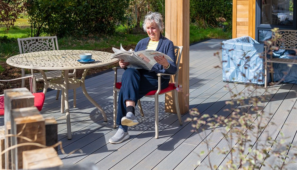 A woman sitting on a garden chair on Composite Decking Boards drinking a cup of a tea and reading a newspaper